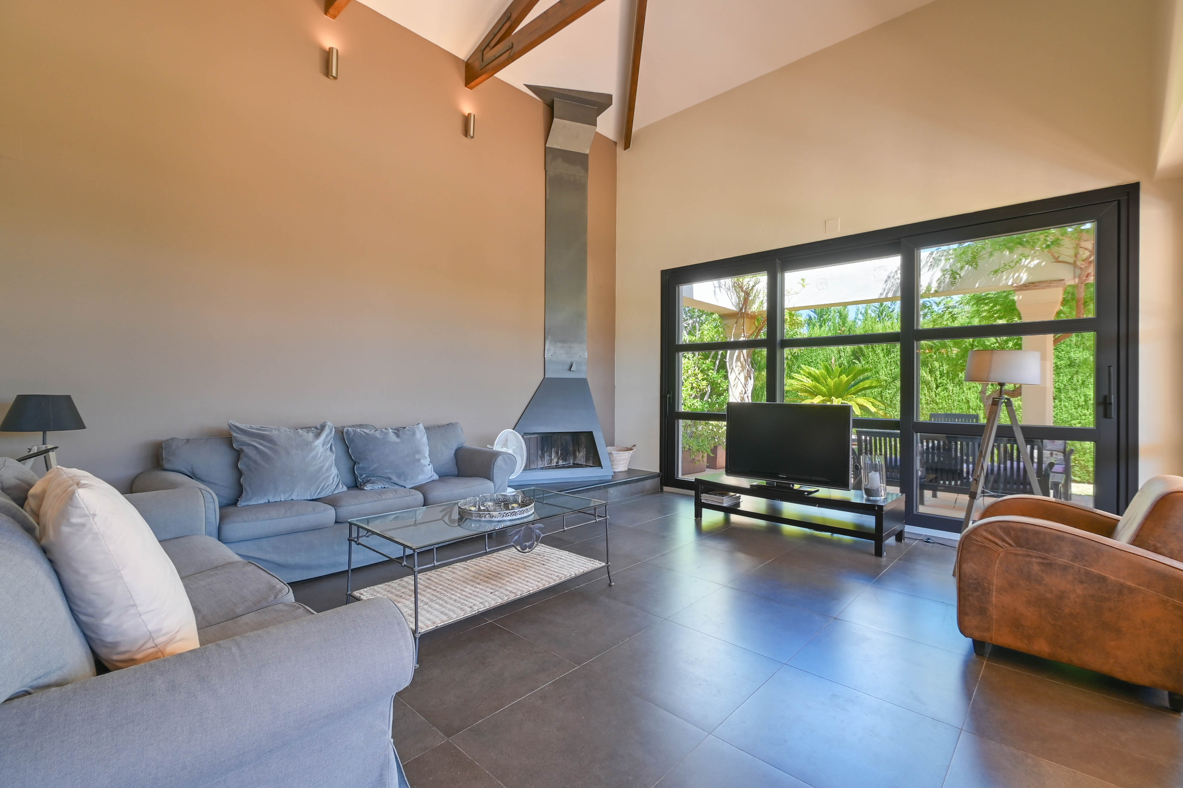 Geräumiges Wohnzimmer mit Metallkamin, grauem Sofa und großen Fenstern mit Blick auf den Garten in Denia, Costa Blanca.