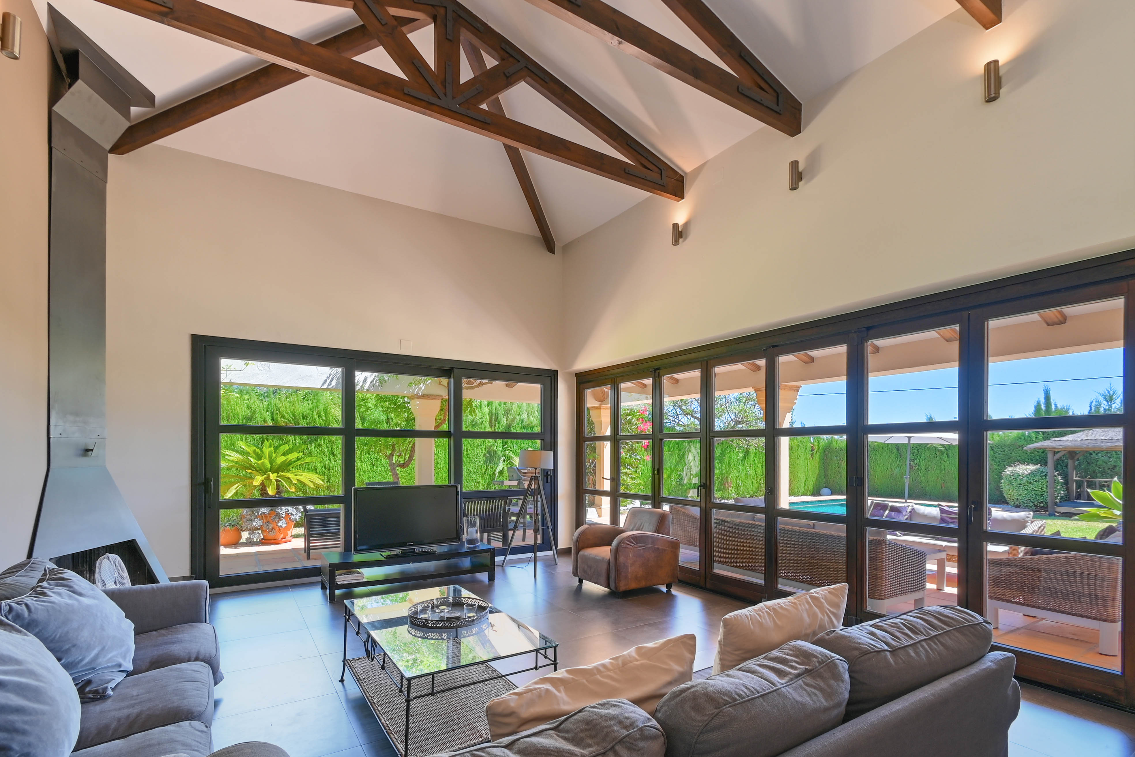 Helles Wohnzimmer mit Holzbalken, modernem Kamin und Gartenblick mit Pool durch große Fenster.