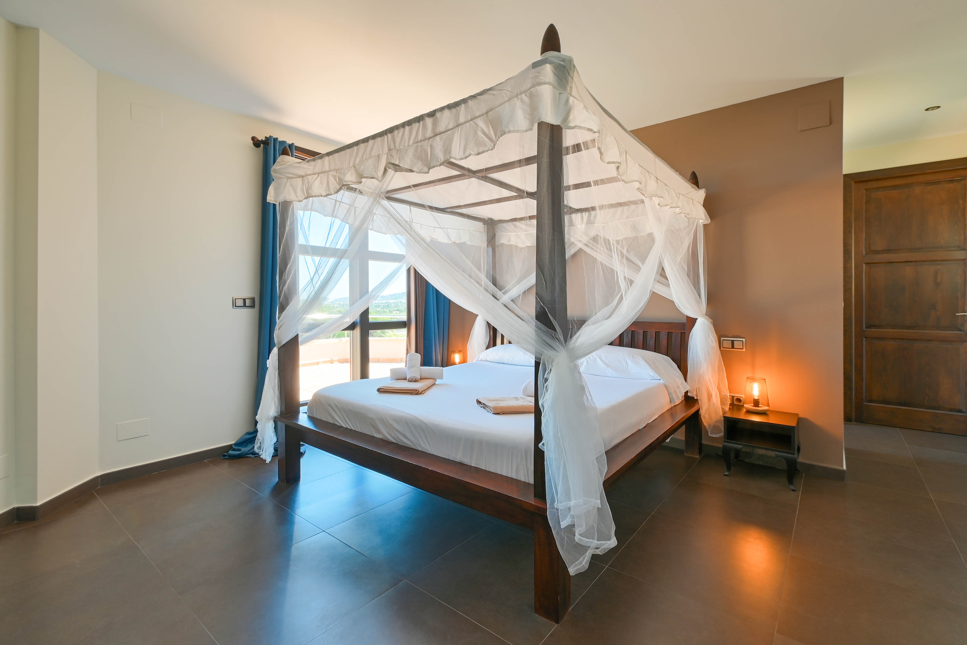 Schlafzimmer mit dunklem Holzhimmelbett mit weißen Vorhängen und braunem Fliesenboden.