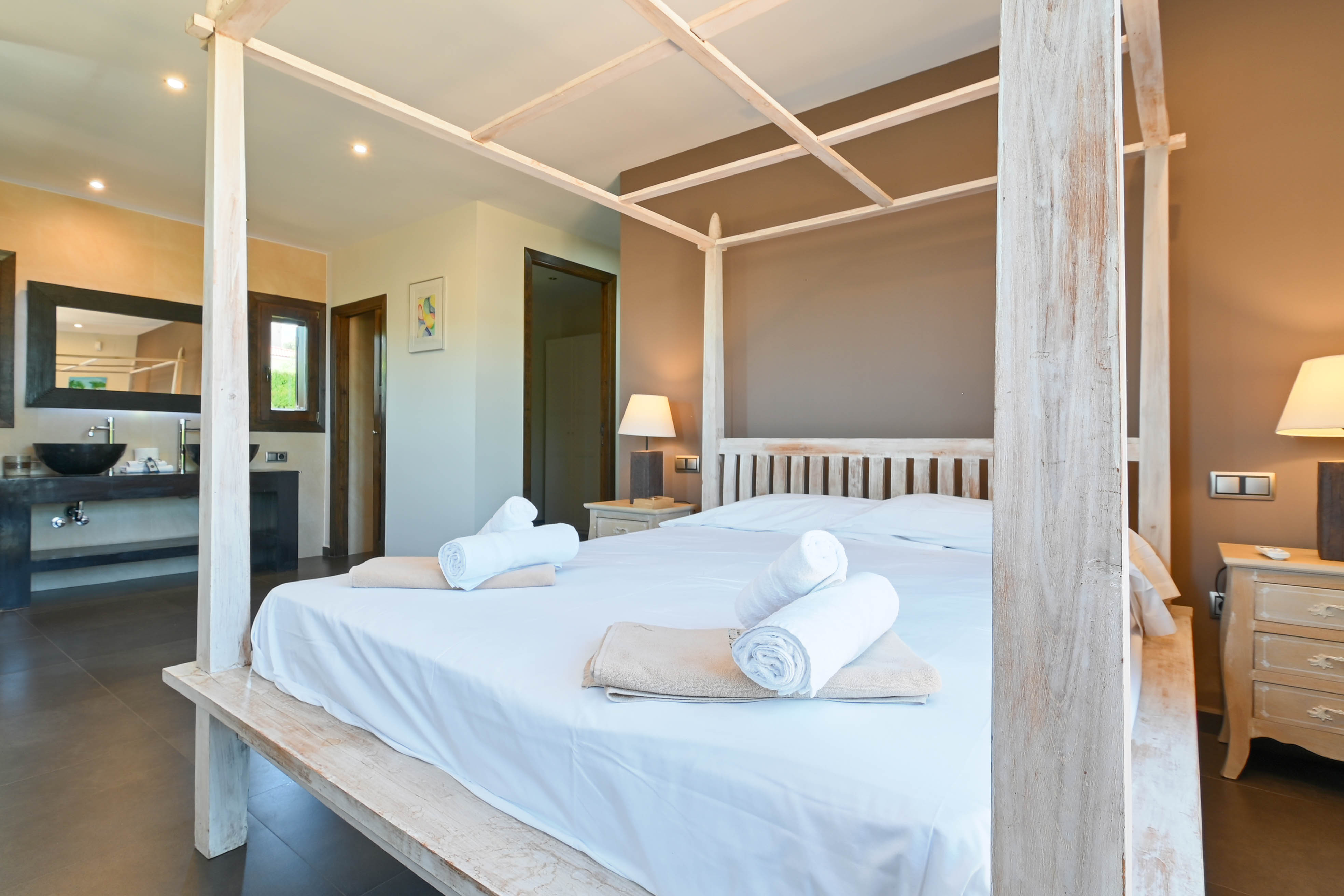 Schlafzimmer mit weißem Holzhimmelbett, gerollten Handtüchern und Badezimmer mit modernem Waschbecken.