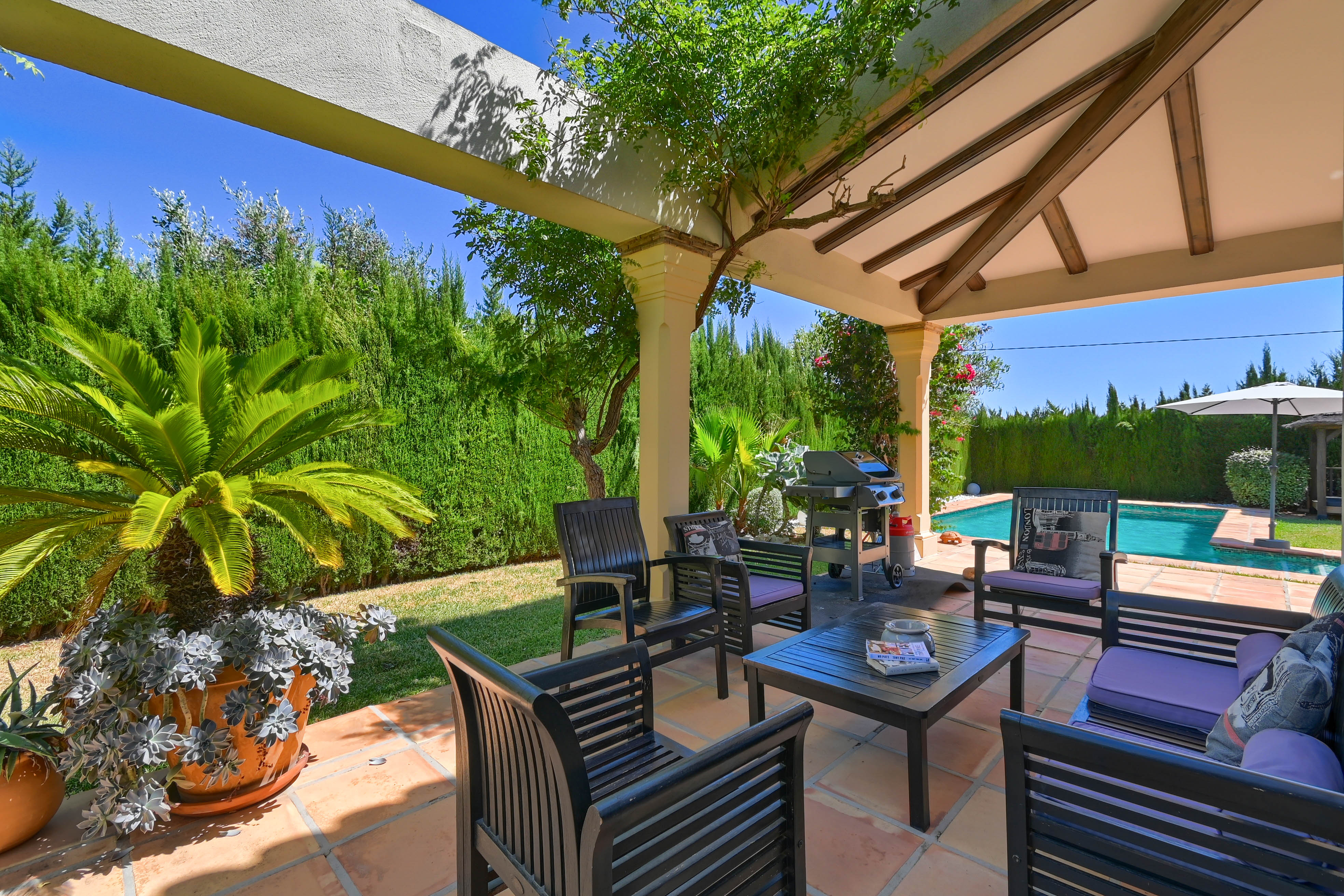 Überdachte Veranda mit dunklen Holzsitzen, lila Kissen mit Blick auf den Pool und den Garten.