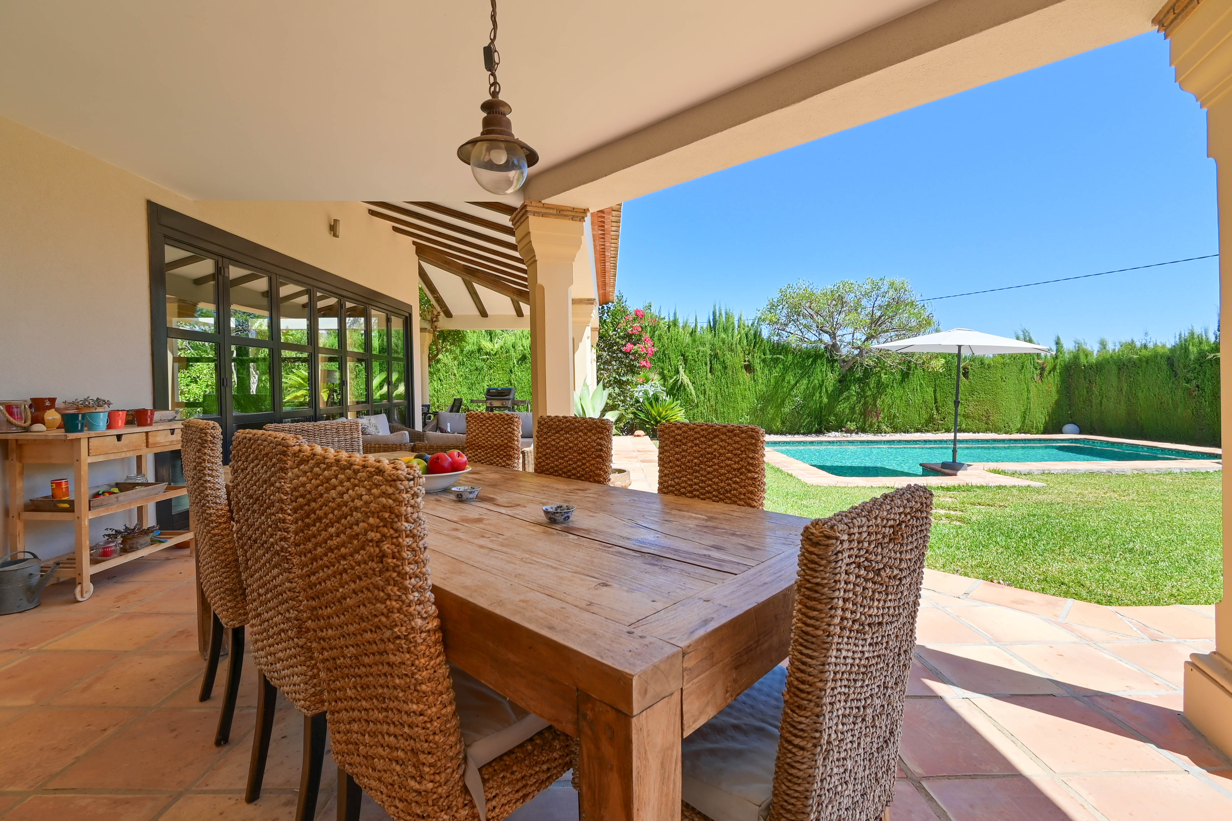 Überdachter Essbereich im Freien mit Massivholztisch und Rattanstühlen mit Blick auf den Pool und den Garten.