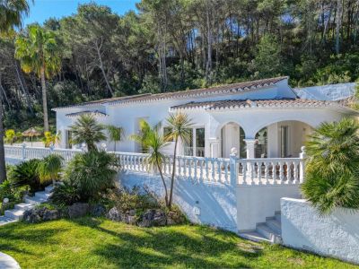 Außenansicht der Villa mit überdachter Terrasse, Balustradengeländer und Garten mit mediterraner Vegetation.