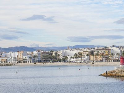 Panoramablick auf den Strand und die Promenade von Jávea, mit Wohngebäuden und Bergen im Hintergrund.