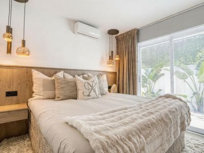 Schlafzimmer mit Holzkopfteil, Glaspendelleuchten und beigefarbenen Vorhängen neben einem Fenster.