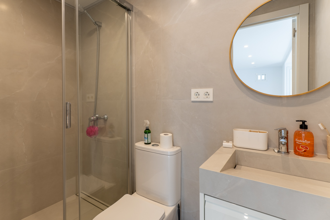 Badezimmerdetail mit Glasdusche, modernem Waschbecken und rundem Spiegel mit goldenem Rahmen.