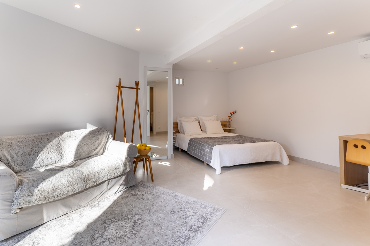 Helles Schlafzimmer mit Sofa, Doppelbett und Schreibtisch, beleuchtet durch Einbauspots.