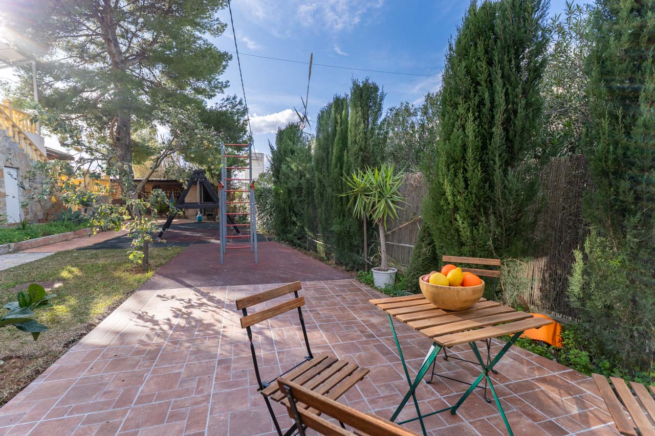 Terrasse mit Holztisch und Obstschale, umgeben von Grün und einem Kinderspielplatz.