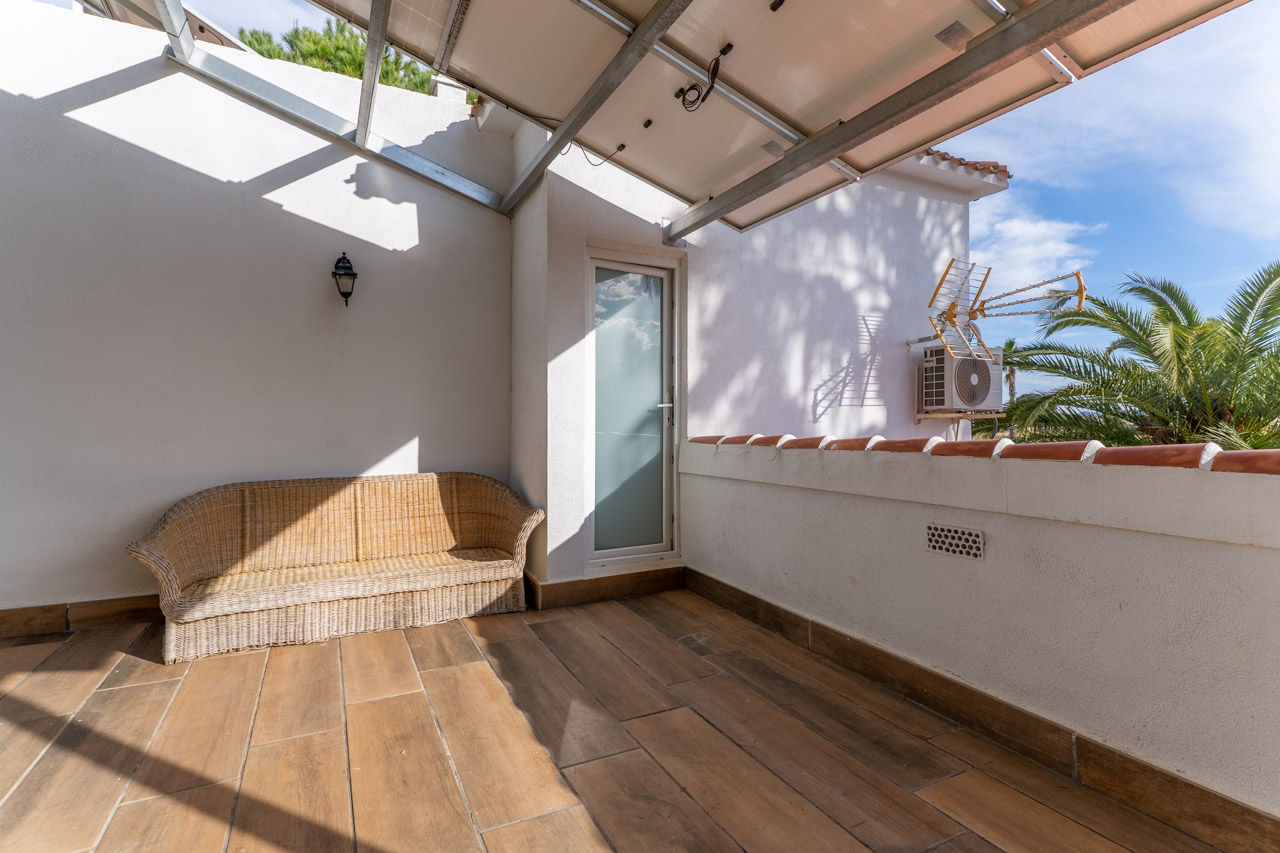 Überdachte Terrasse mit Keramikboden und Korbsofa, beleuchtet durch Tageslicht und mit Zugang nach draußen.