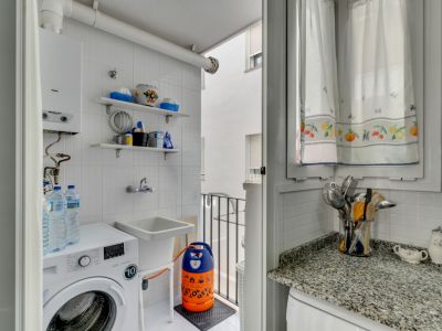 Waschküche mit Waschmaschine, Spülbecken und weißen Fliesenregalen. Fenster mit gemusterten Vorhängen.