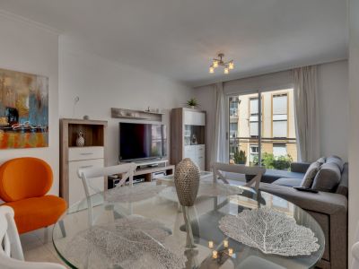 Wohn-Esszimmer mit Glastisch, weißen Stühlen und grauem Sofa, beleuchtet durch Tageslicht.