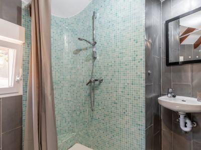 Badezimmerdetail mit türkisfarbener Mosaikdusche, wandmontiertem Waschbecken und grauen Fliesenwänden.
