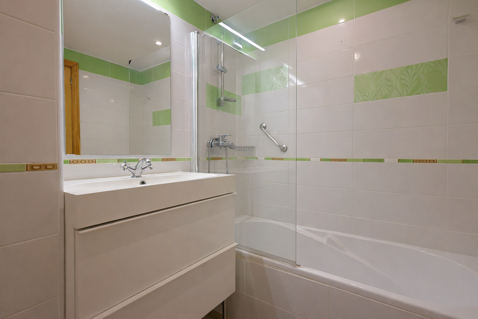 Badezimmer mit modernem Waschbecken, weißen Fliesen mit grünen Akzenten und gläserner Duschkabine.