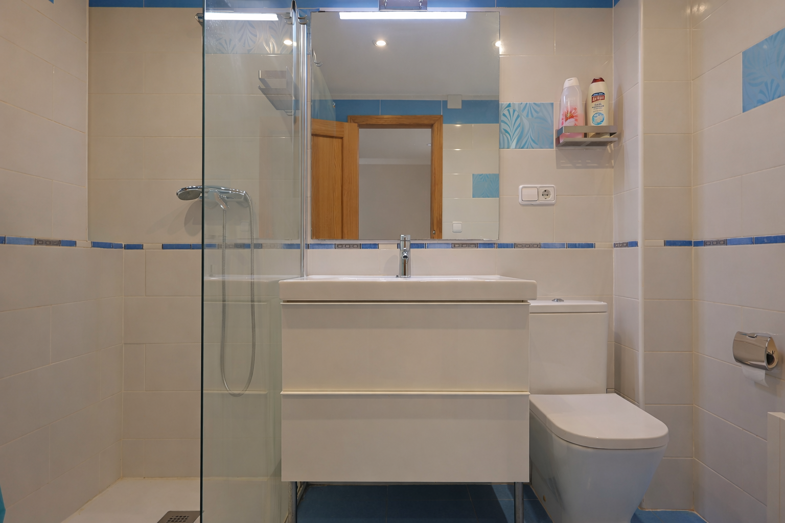 Badezimmer mit weißen Fliesen, blauen Akzenten, Spiegel, modernem Waschtisch und Glasdusche.