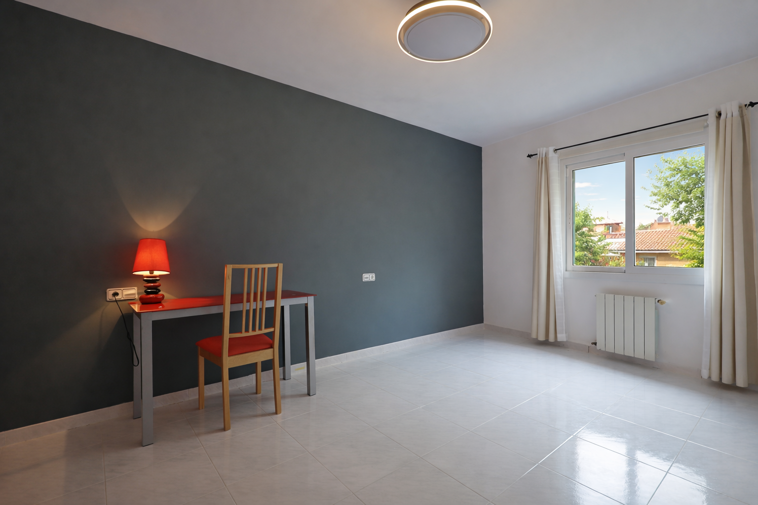 Helles Zimmer mit rotem Schreibtisch und bleigrauer Wand, cremefarbenen Bodenfliesen und runder Deckenleuchte.