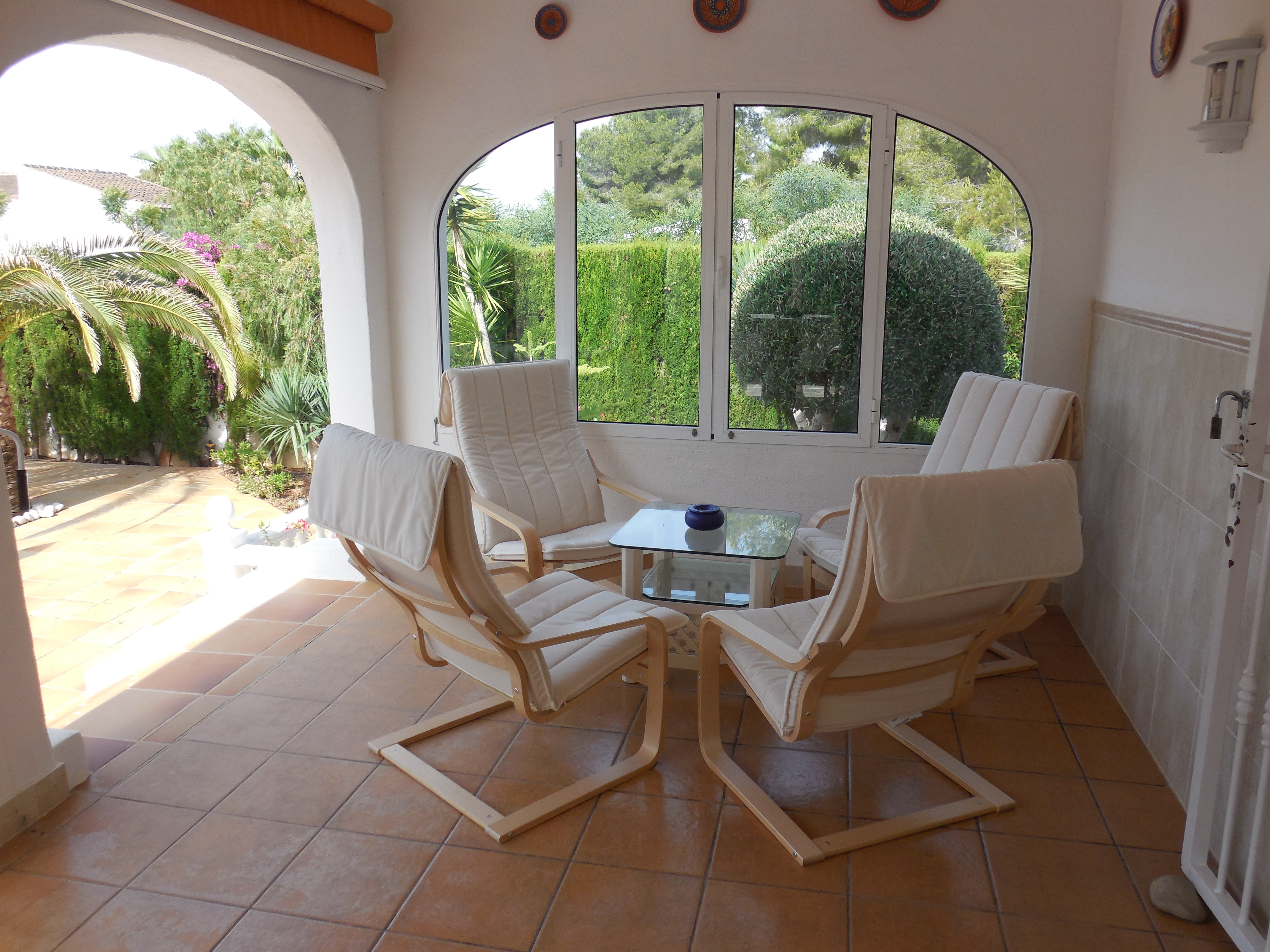Überdachte Terrasse mit vier hellen Holzsesseln mit weißen Kissen, einem Glastisch und Gartenblick.
