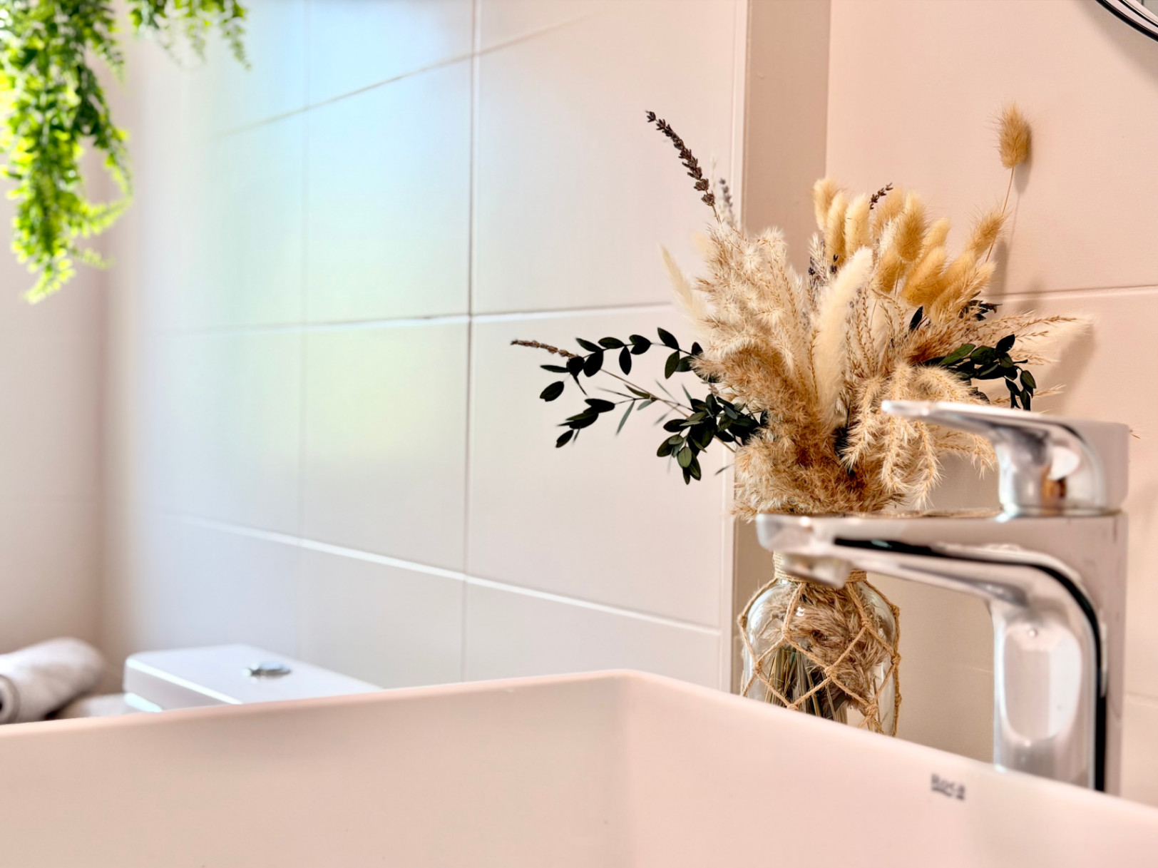 Detail eines renovierten Badezimmers mit moderner Chromarmatur und Trockenblumengesteck auf einer weißen Arbeitsplatte.
