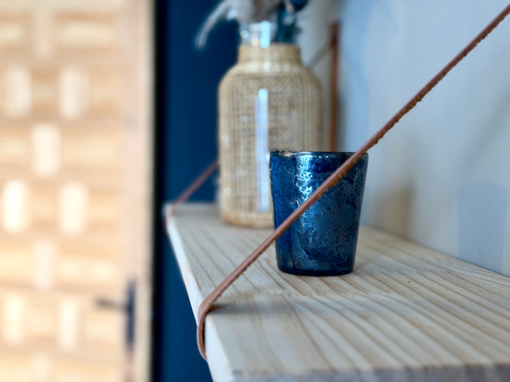 Detailaufnahme eines hellen Holzregals mit Dekoration: ein strukturiertes blaues Glas und eine Glasflasche mit einer Weidenhülse.