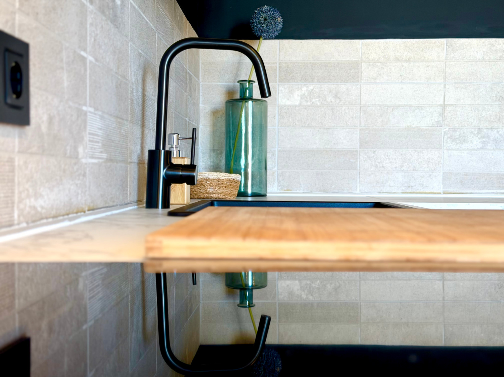 Küchendetail mit schwarzem Wasserhahn, Holzschneidebrett und strukturierter Keramikfliesen-Rückwand.