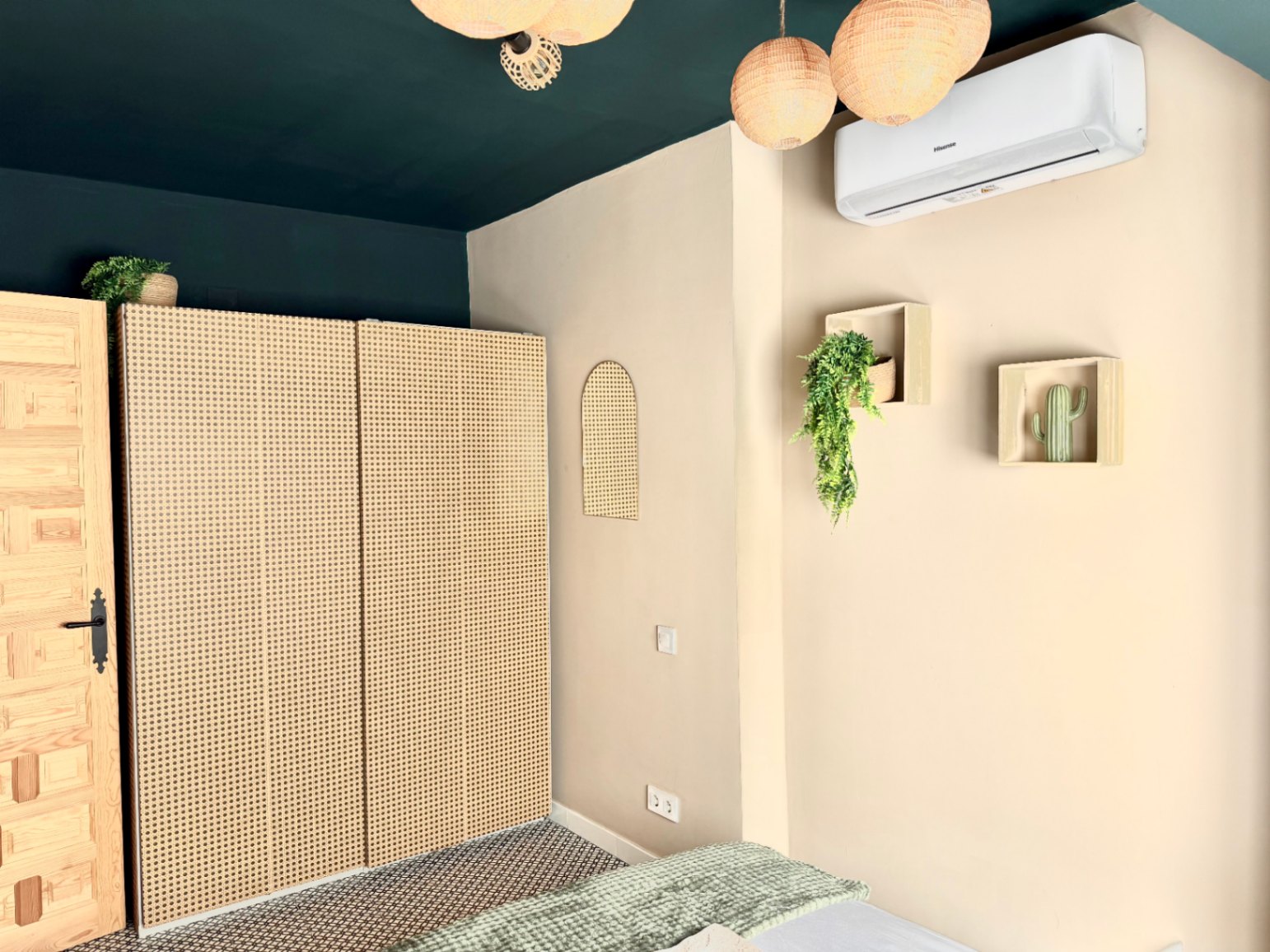 Schlafzimmer mit Rattan-Kleiderschrank, dunkelgrüner Decke und minimalistischer Wanddeko.