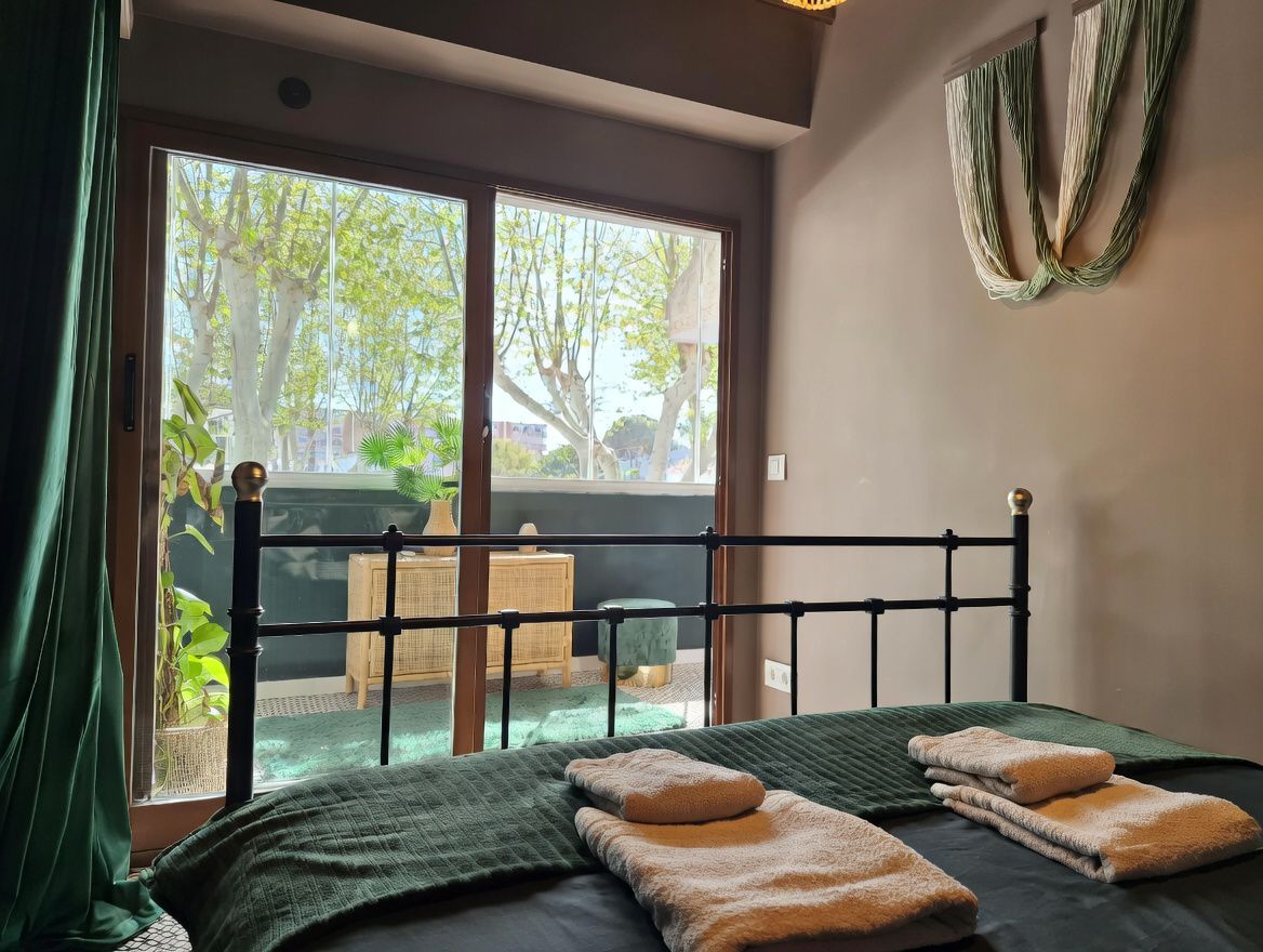 Schlafzimmer mit schmiedeeisernem Bett und Terrasse mit Blick auf Bäume. Minimalistische Einrichtung in neutralen und Grüntönen.