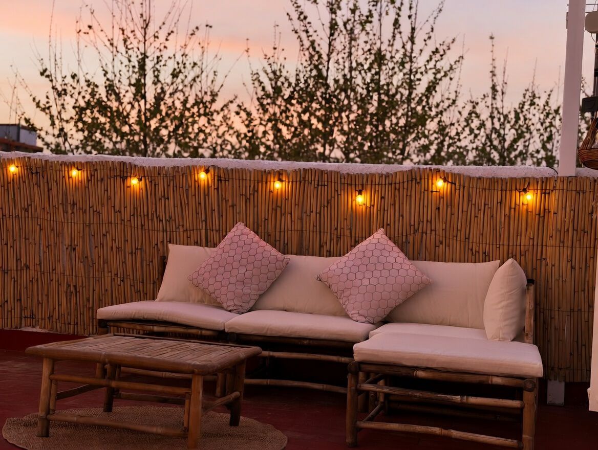 Terrasse mit Bambussofa und gemusterten Kissen, beleuchtet mit warmen Lichterketten, mit Blick auf den Sonnenuntergang.