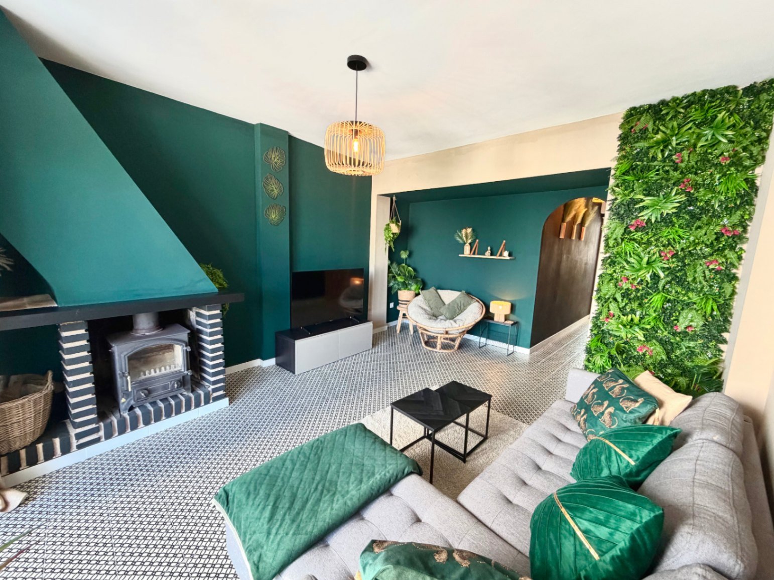 Wohnzimmer mit rustikalem Kamin, grüner Wand, geometrischen Bodenfliesen und einer Hängelampe aus Korbgeflecht.