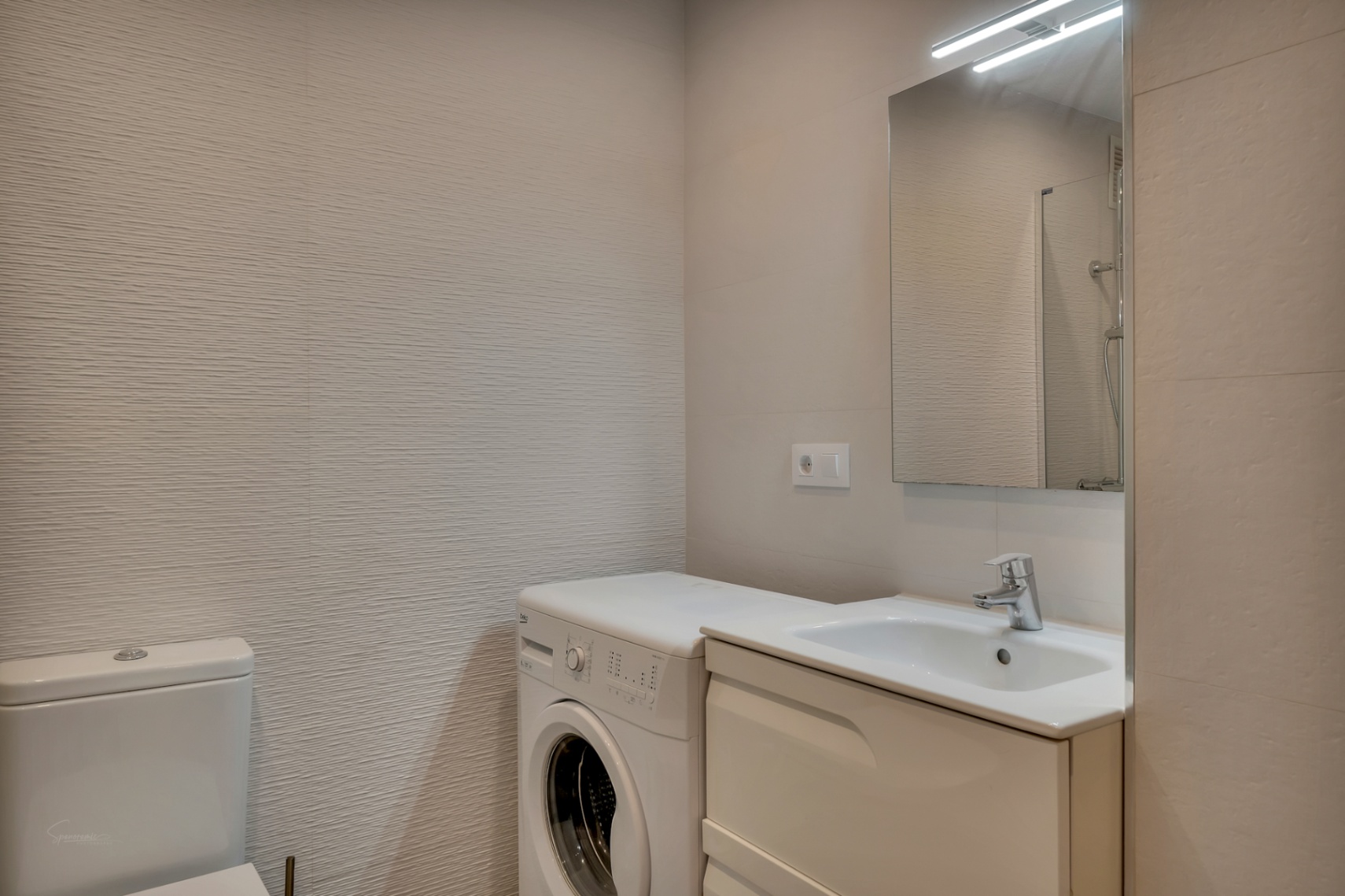 Badezimmerdetail mit Einbauspüle, Waschmaschine und strukturierten Wänden in neutralen Tönen.