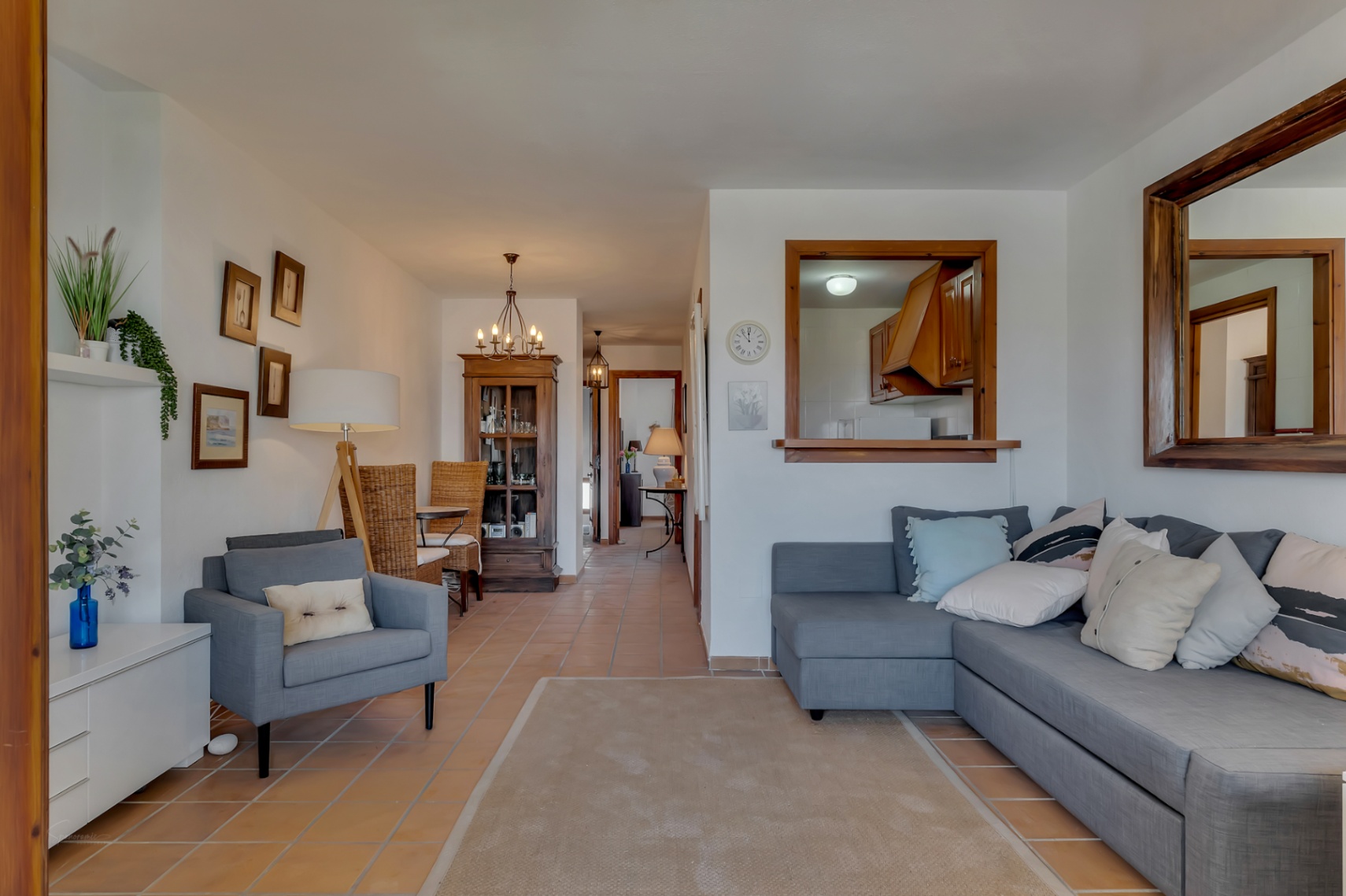 Wohnzimmer mit Terrakottaboden, grauem Sofa und Blick in die Küche durch ein Innenfenster in Jávea.