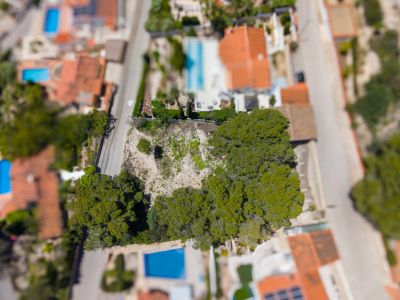 Luftaufnahme eines unbebauten Grundstücks mit Bäumen, umgeben von Straßen und Wohnhäusern mit Pools und roten Dächern.