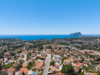 Panoramische Luftaufnahme der Küste von Benissa, mit dem Mittelmeer, dem Peñón de Ifach und einer Wohngegend mit üppiger Vegetation unter klarem blauem Himmel.