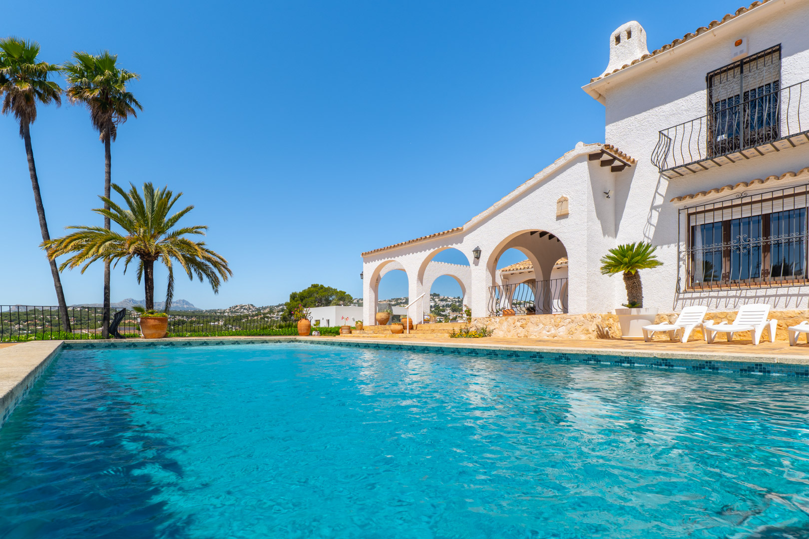 Türkisfarbener Pool mit Mosaikrand und weiße Villa im mediterranen Stil mit Bögen.