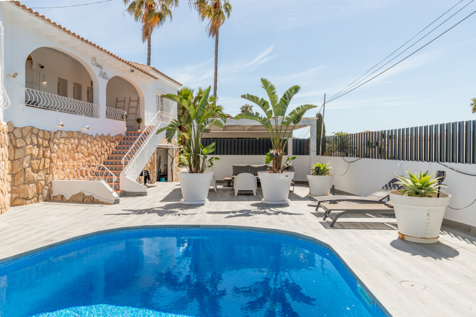 Außenansicht der Villa 'Casa Alta' mit blauem Mosaikpool. Die helle Holzterrasse umgibt den Pool, angrenzend an eine Außentreppe mit dekorativen Fliesen und Steinsockel.