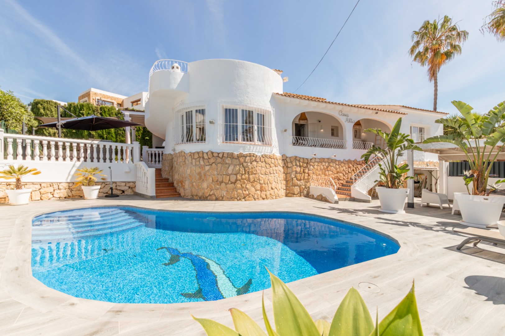 Außenansicht einer weißen Villa mit privatem blau gefliestem Pool und Delfinmosaik. Steinfassade und ein markanter runder Gebäudeteil sind sichtbar.