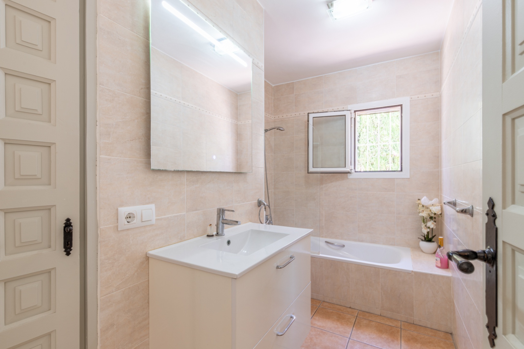 Badezimmer mit hellbeigen Fliesen, weißem Waschtischunterschrank und großem Spiegel mit Beleuchtung. Ausgestattet mit Badewanne und Fenster für Tageslicht.
