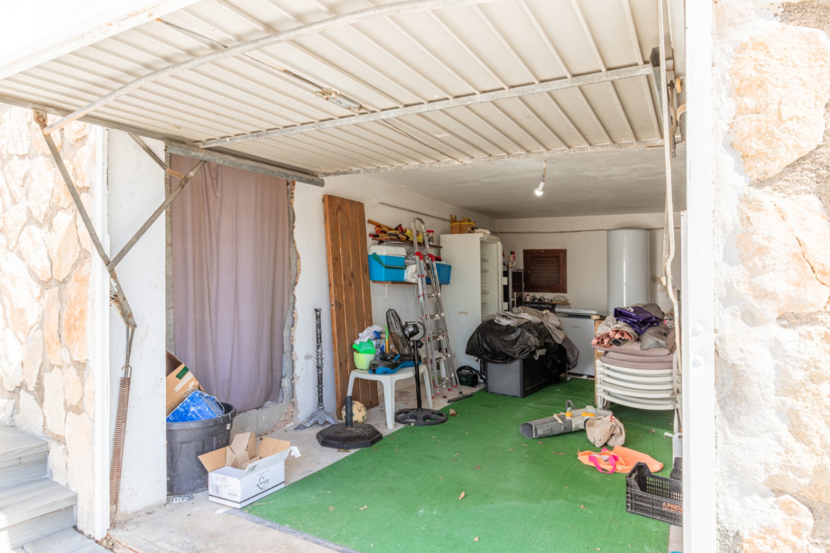 Garage oder Abstellraum mit Wellblechdach und grünem Kunstrasenboden. Enthält einen Warmwasserbereiter, Kühlschrank, Leiter und diverse Lagergegenstände.