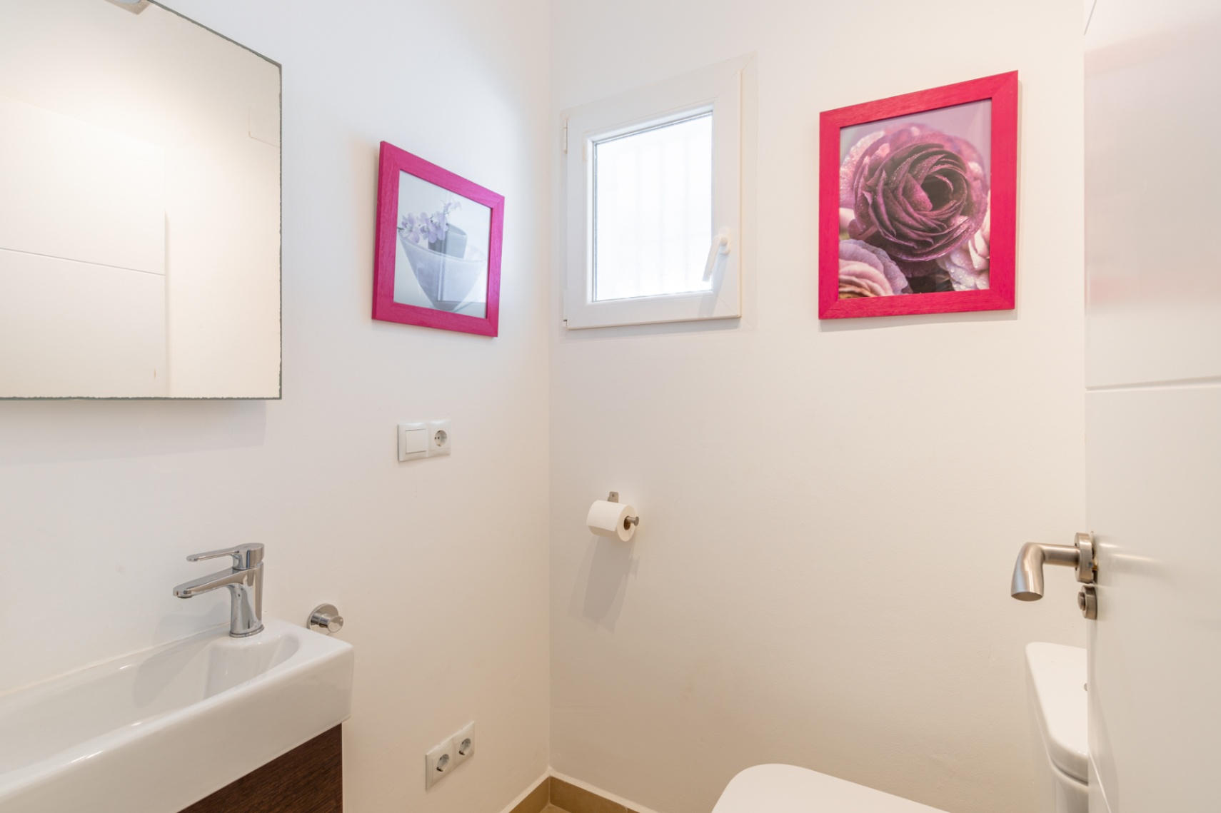 Gäste-WC mit rechteckigem Waschbecken, modernem Chromhahn und dunklem Holzwaschtisch. Ein kleines Fenster sorgt für Tageslicht, ergänzt durch Bilder mit pinken Rahmen.