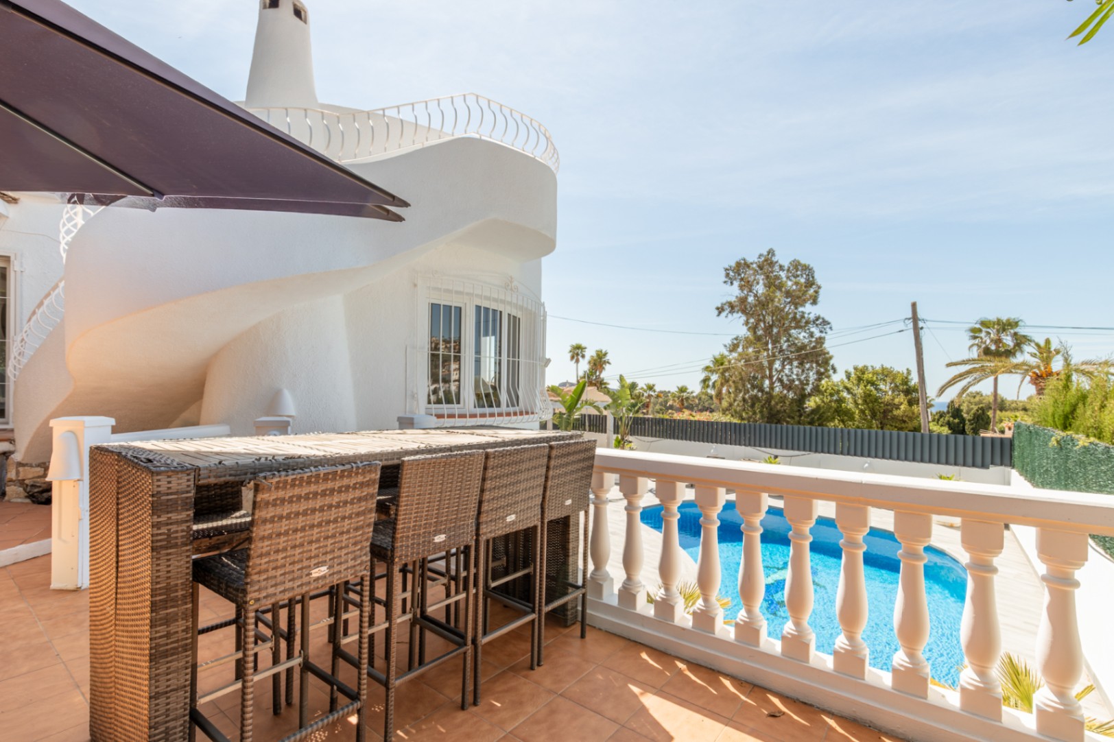Geflieste Außenterrasse mit Rattan-Bartisch und Hockern, mit Blick auf den privaten Pool. Die weiße Villa zeigt ein einzigartiges spiralförmiges Architekturelement bei klarem Himmel.