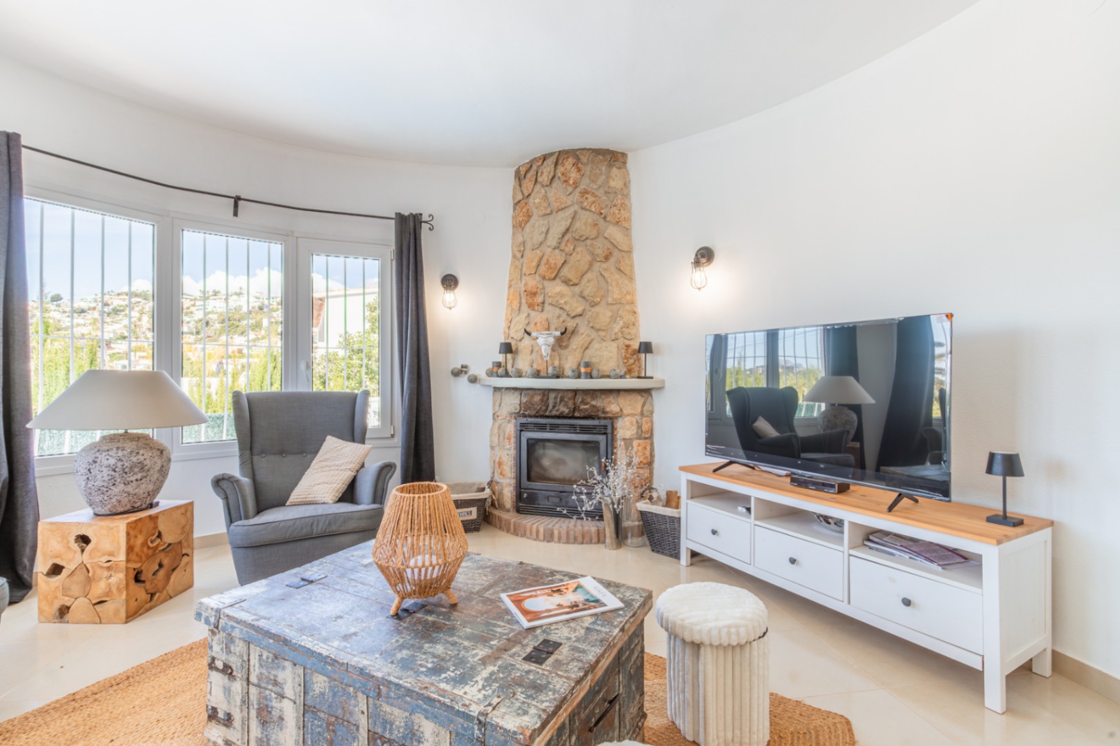 Gemütliches Wohnzimmer mit steingemauertem Kamin und großen Erkerfenstern mit Blick auf die Hügel. Ein rustikaler Couchtisch und ein modernes TV-Möbel ergänzen den Raum.