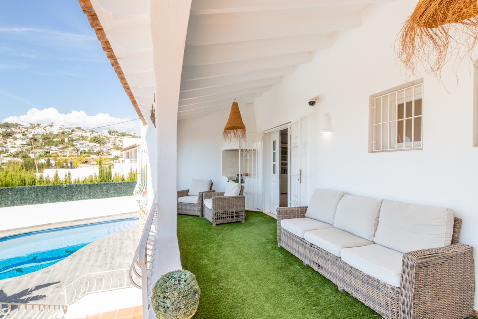 Geräumige überdachte Terrasse mit gemütlichen Korbmöbeln auf Kunstrasen, bietet Blick auf den privaten Pool und die bewachsenen Hügel in der Ferne.