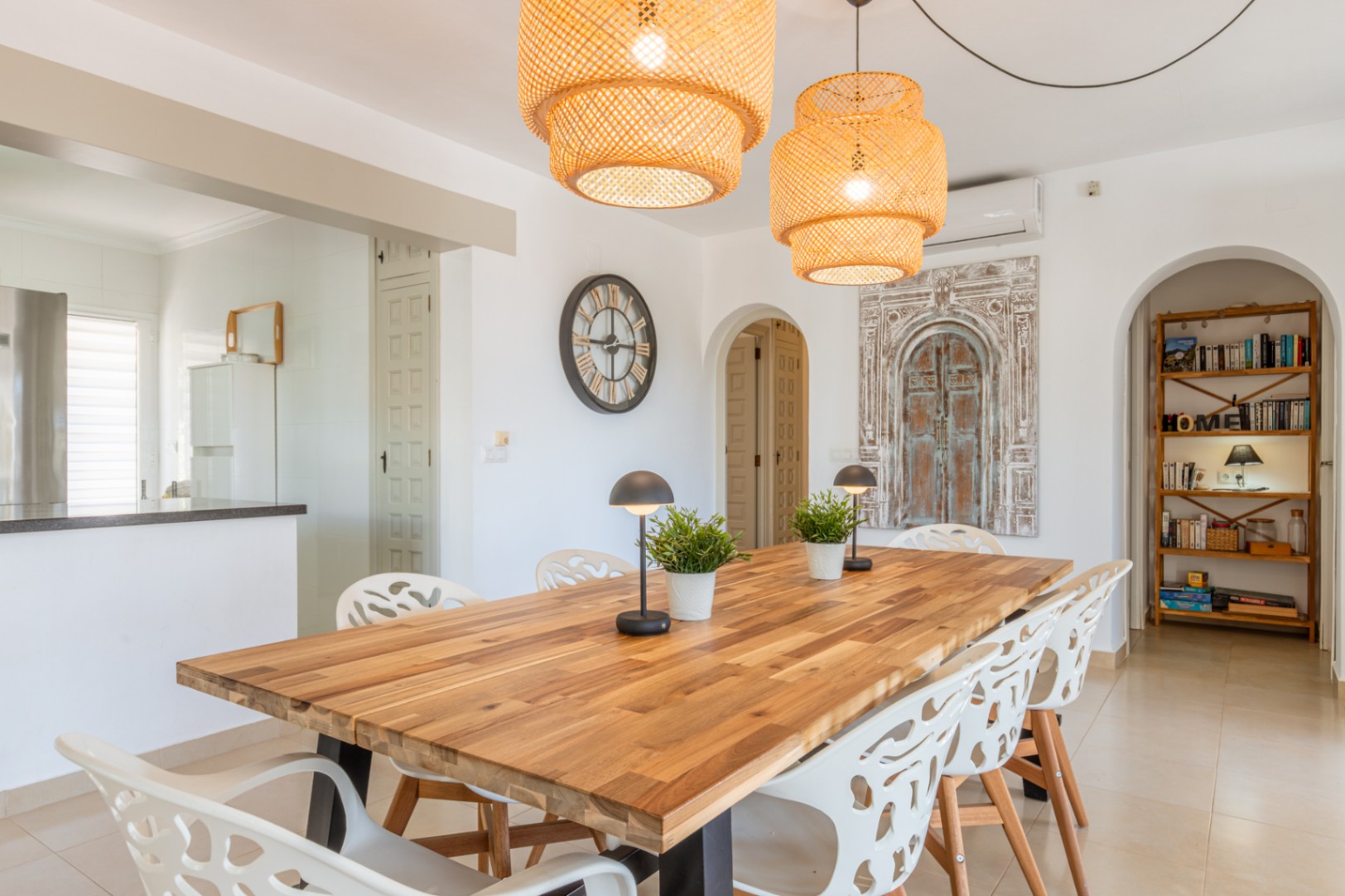 Geräumiger Essbereich mit Holztisch, modernen weißen Stühlen und zwei geflochtenen Hängelampen. Bögen führen zur Küche und einem Lesebereich mit Holzregal.