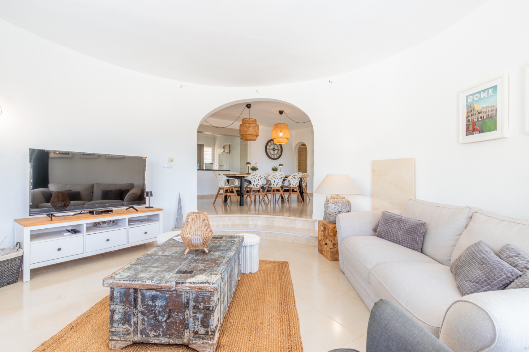 Geräumiges Wohnzimmer mit hellen Fliesen, bequemen Sofa und rustikalem Holztisch auf Jute-Teppich. Ein Bogen führt zum erhöhten Essbereich mit modernen Stühlen und Rattan-Hängeleuchten.
