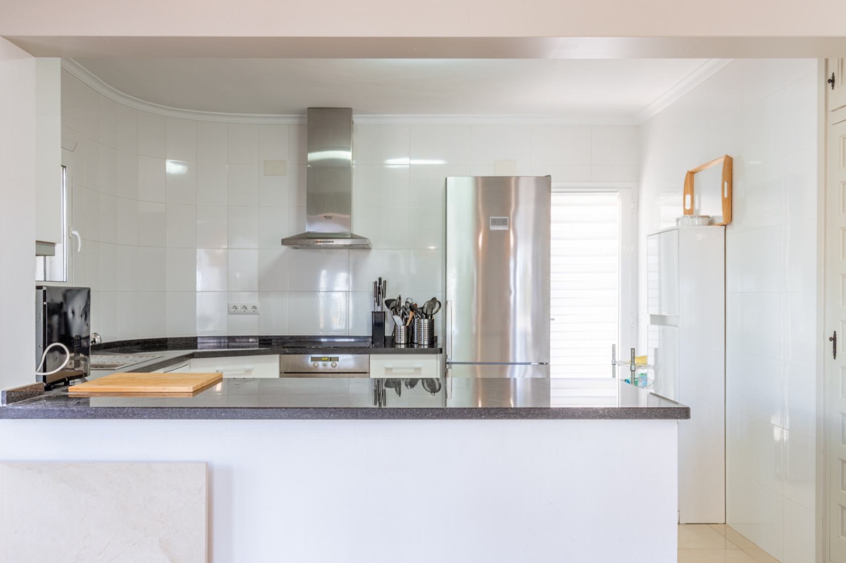 Helle, moderne Küche mit weißen Fliesenwänden, dunklen Granitarbeitsplatten und integrierten Edelstahlgeräten wie Kühlschrank und Dunstabzugshaube. Eine offene Theke bietet Arbeitsfläche.