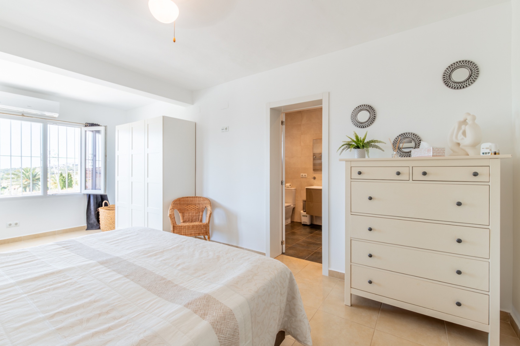 Helles Hauptschlafzimmer mit Doppelbett, weißem Kleiderschrank und direktem Zugang zum eigenen Bad. Das Fenster bietet Blick auf die Außenvegetation.