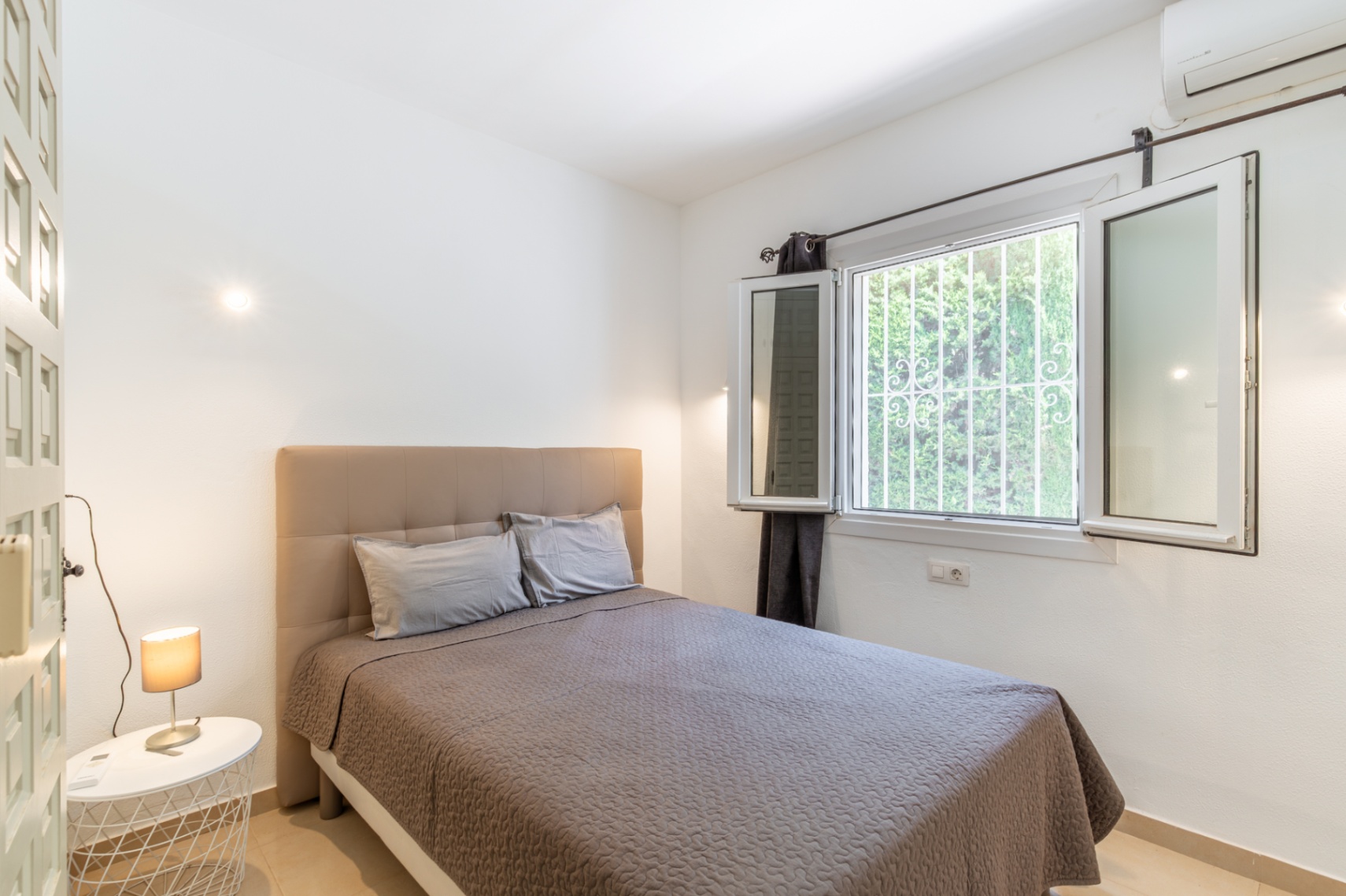 Helles Schlafzimmer mit bequemem Bett und gepolstertem Kopfteil, großem Fenster mit Sicherheitsgittern und Blick ins Grüne, sowie Klimaanlage.