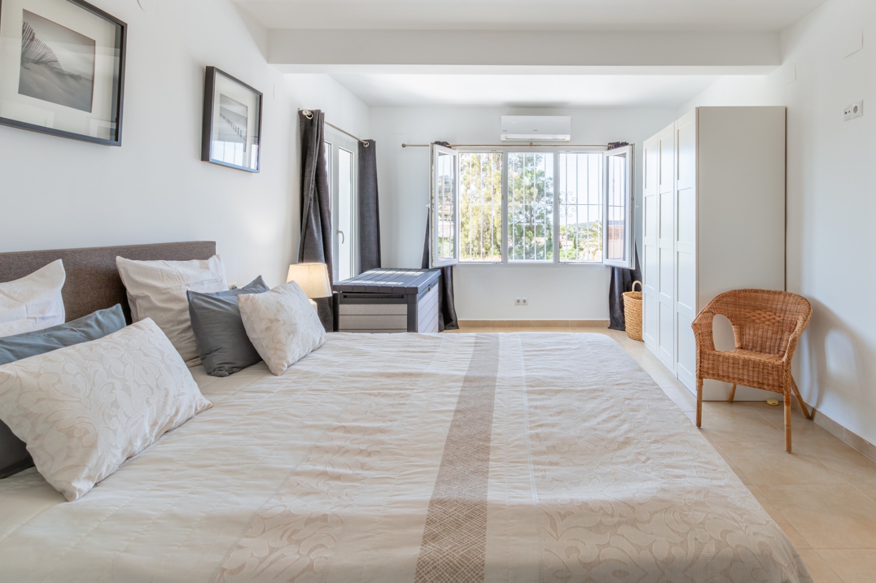 Helles Schlafzimmer mit großem Bett, hellen Fliesen, weißem Kleiderschrank und vergittertem Fenster mit Blick ins Grüne, inklusive Klimaanlage.