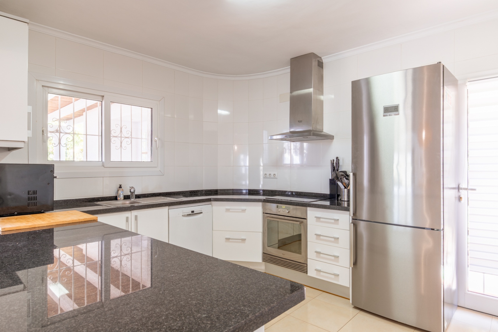 Küche mit weißen Schränken, dunklen Granitarbeitsplatten und Edelstahlgeräten wie Kühlschrank und Einbaubackofen. Fenster mit Sicherheitsgittern bietet Tageslicht.