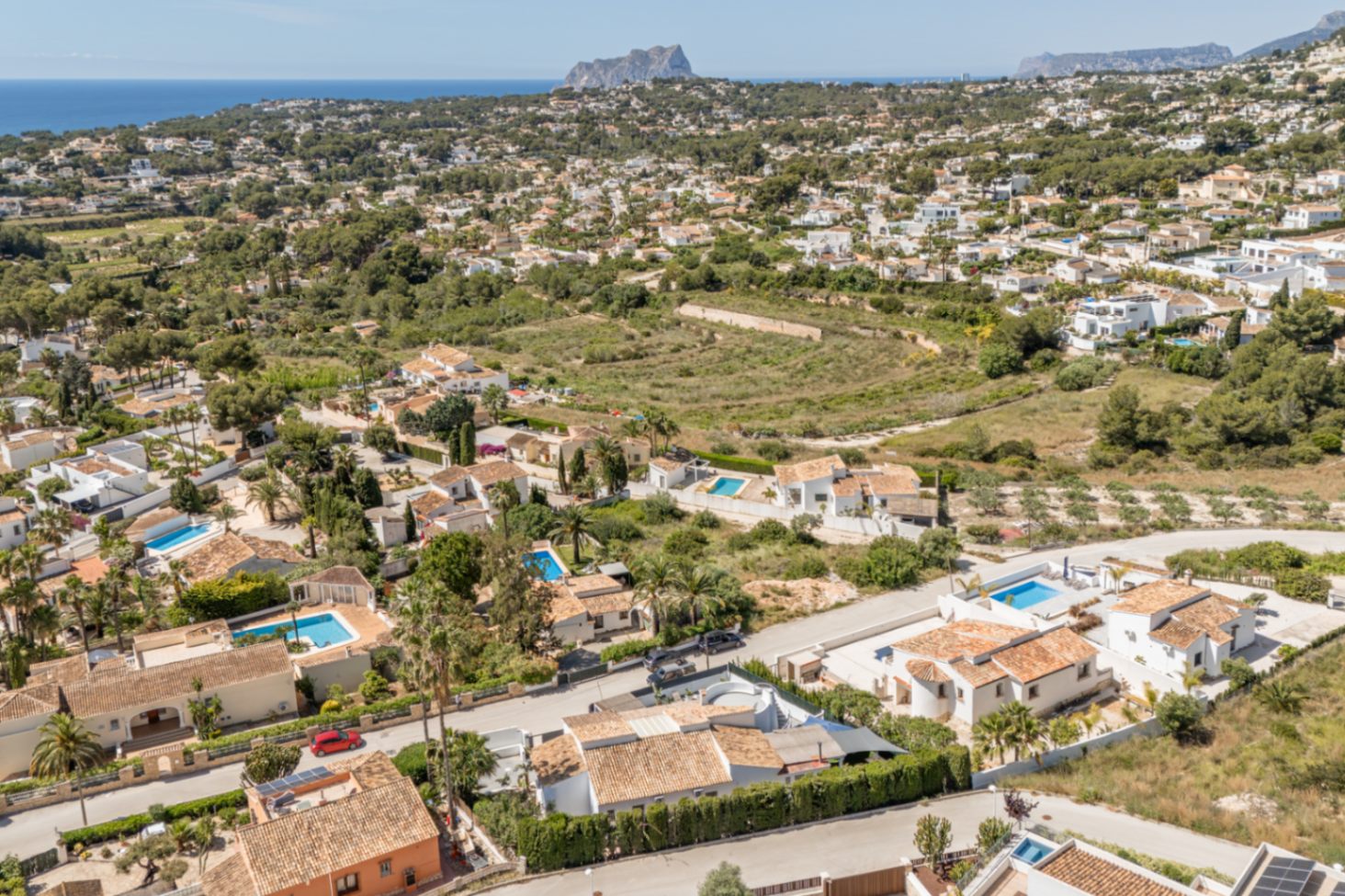Luftaufnahme eines Wohngebiets in Moraira, mit zahlreichen Villen mit Terrakottadächern, privaten Pools und üppiger Vegetation. Das Mittelmeer und eine ferne Felsformation prägen die Küstenlandschaft.