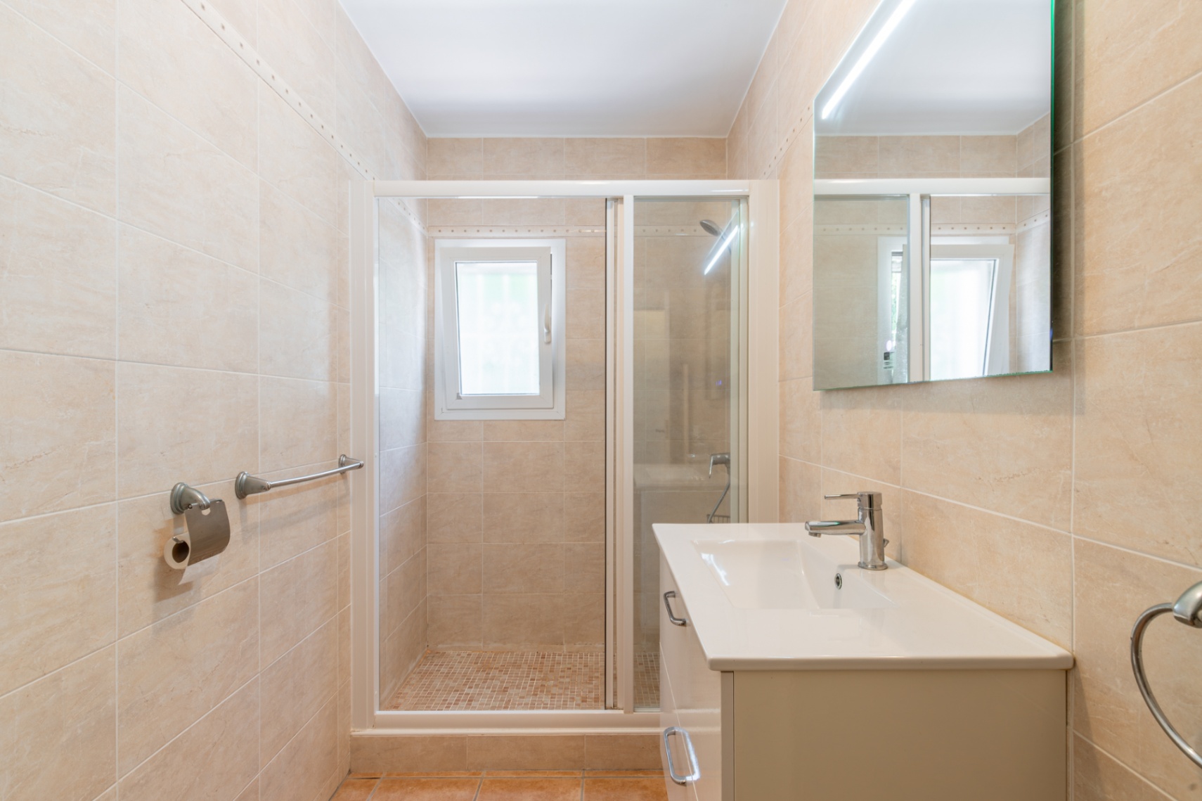 Modernes Badezimmer mit beigen Wandfliesen, weißem Waschtisch, LED-Spiegel und einer begehbaren Dusche mit Mosaikboden und Fenster.
