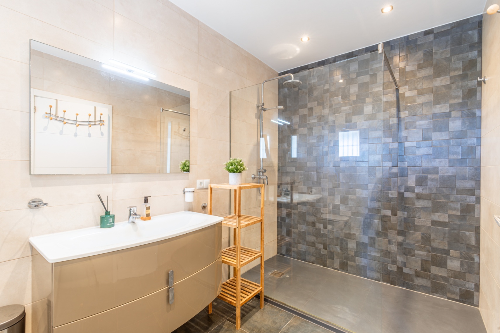 Modernes Badezimmer mit glänzend beigem Waschtisch, beleuchtetem Spiegel und ebenerdiger Dusche mit Glaswand und dunkelgrauen Mosaikfliesen. Helle Wandfliesen, dunkler Boden.